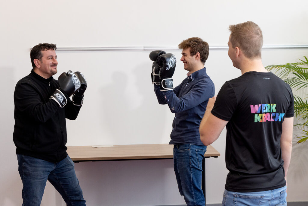 Rechts staat werkcoach Sjoerd van Eck, hij heeft kort, blond haar en draagt een spijkerbroek en zwart shirt met gekleurd Werkkracht-logo. Hij coacht twee mannen die allebei bokshandschoenen dragen en lachend tegenover elkaar staan.