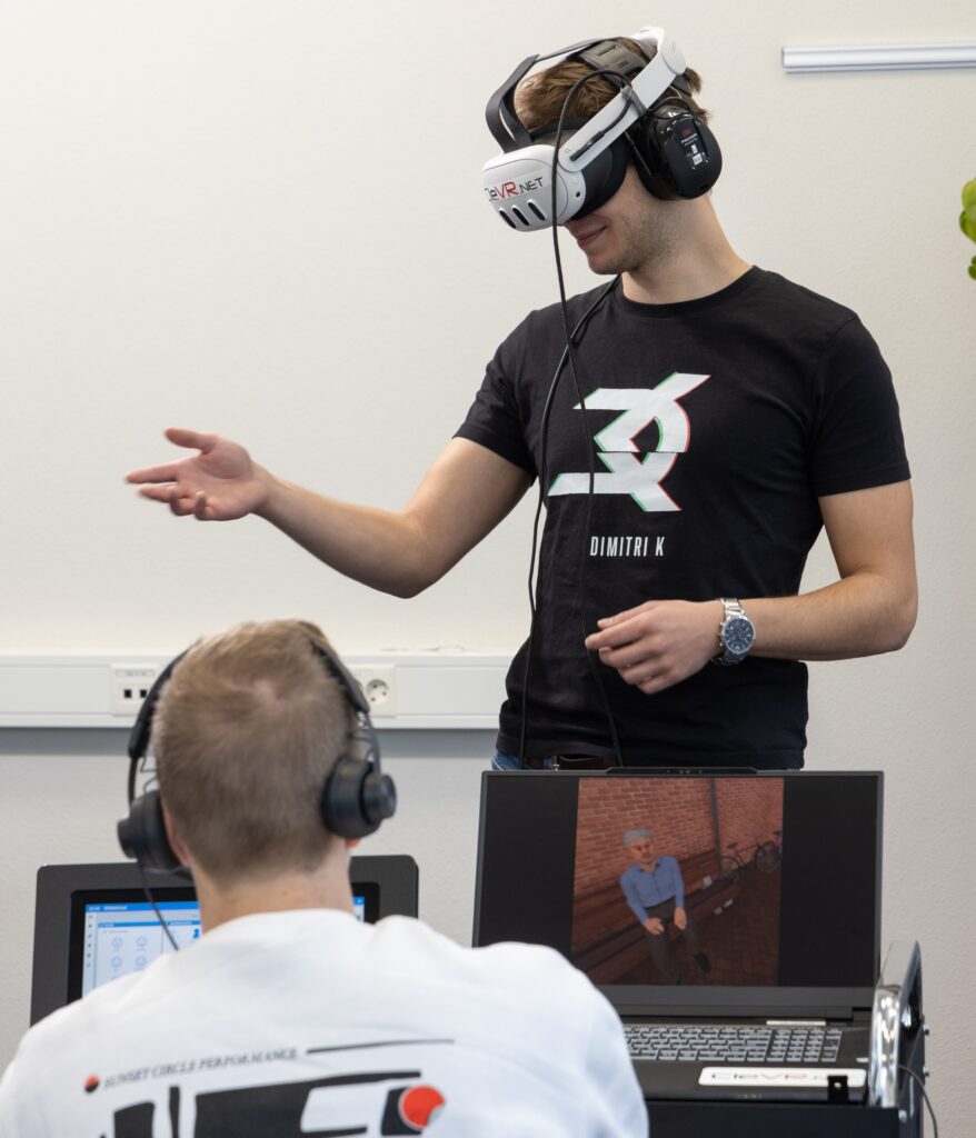 Een man draagt een witte VR-bril en een zwarte koptelefoon. Hij draagt een zwart shirt met een witte afbeelding en strekt zijn rechterarm uit alsof hij iemand een hand geeft. Op de voorgrond zit werkcoach Sjoerd van Eck in een witte trui. Ook hij draagt een koptelefoon, en kijkt naar twee laptops, op een van de laptops is een virtuele persoon te zien die op een houten bankje zit.