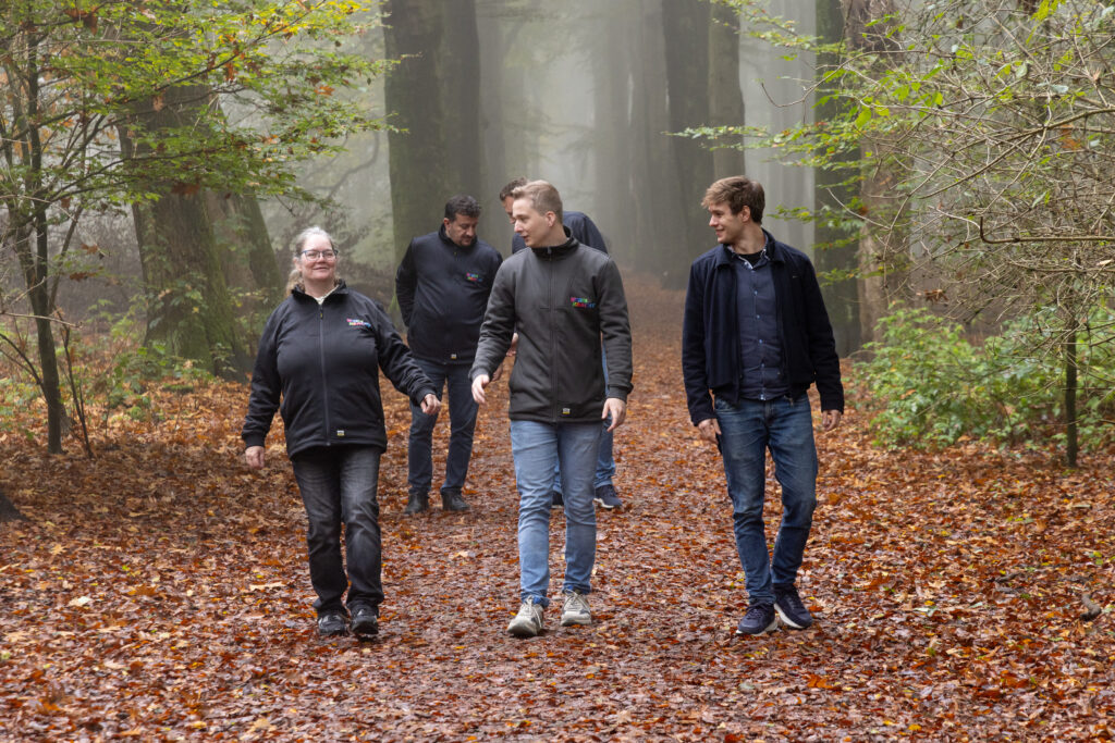 Op de achtergrond zijn lange, hoge boomstammen te zien, verhuld in mist. Wat jonge boompjes met groene bladeren staan aan de zijkanten. Op de grond van het bos liggen bruine bladeren. In het midden lopen drie mensen: links een vrouw met een bril, een zwarte spijkerbroek en een zwart vest. In het midden werkcoach Sjoerd van Eck met een spijkerbroek en grijs vest. Rechts loopt een man in spijkerbroek, met een blauw overhemd en daarover een zwart vest. Achter het drietal lopen twee mannen die met elkaar overleggen.
