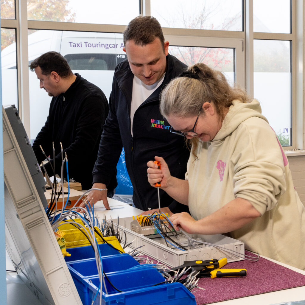 Werkcoach Marius Cornelissen staat naast een kandidaat. Zij heeft grijs haar in een staart en draagt een beige hoodie en heeft een bril. Ze houdt een oranje schroevendraaier vast en draait iets aan in een bakje waar kabels uit lopen. Marius heeft kort bruin haar en draagt een wit t-shirt met daaroverheen een zwart Werkkracht-vest. Op de achtergrond is een andere kandidaat aan het werk, hij draagt een zwarte trui.